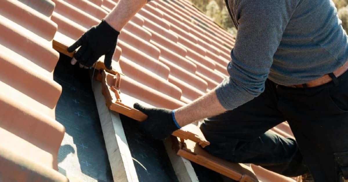 Een dakwerker in zwarte broek en grijze trui legt nieuwe dakpannen op de pannenlatten van een gerenoveerd dak.