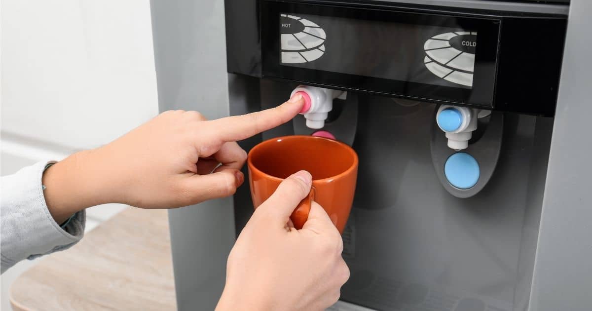 Een dame houdt een rode kop onder de warme kraan van een leidingwaterdispenser in de keuken van een kantoor.