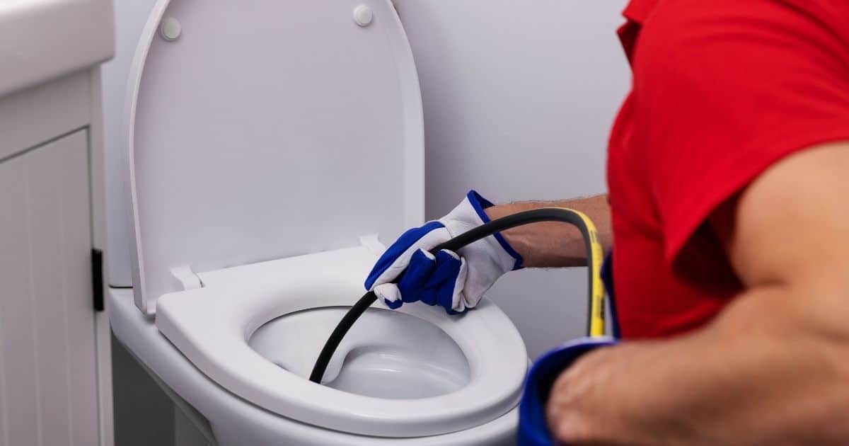 Een ontstoppingsspecialist doet een camera-inspectie in een verstopt toilet. Hij draagt een rood t-shirt en wit met blauwe werkhandschoenen terwijl hij de flexibele camerabuis inbrengt in het witte toilet.