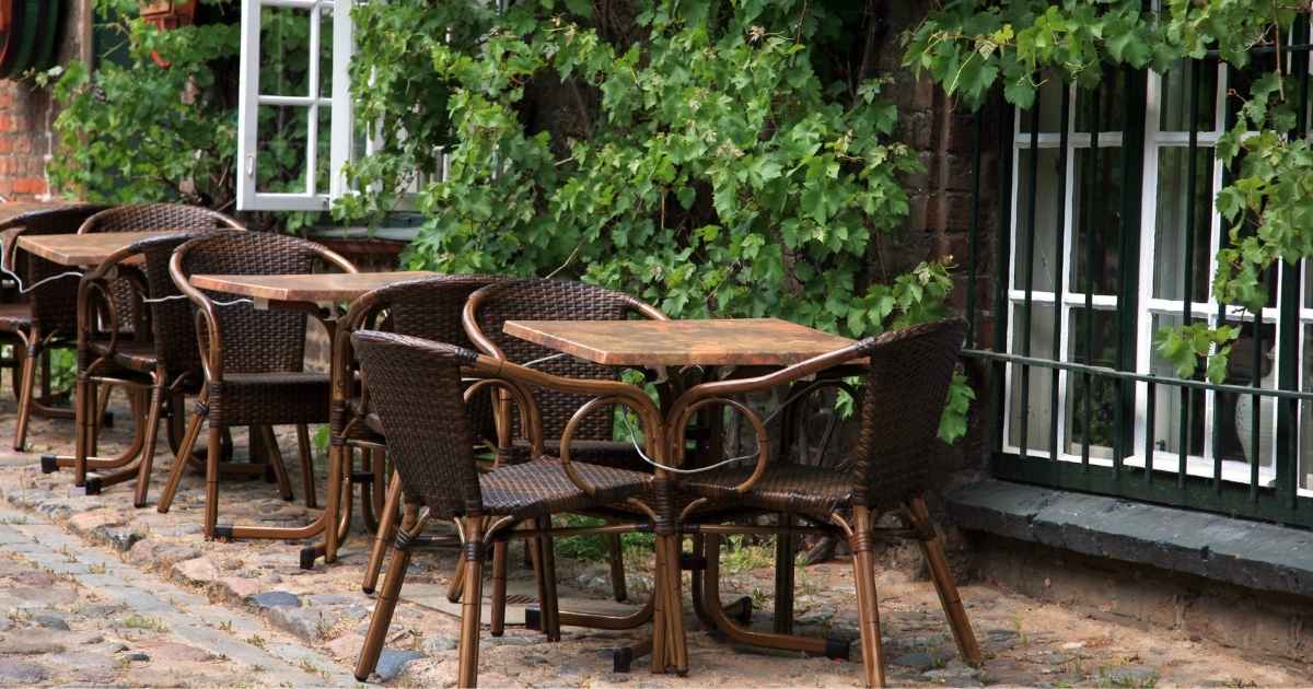 Drie tafels met elks drie stoelen op het terras van een horecazaak. Tegen de gevel groeit een klimplant.
