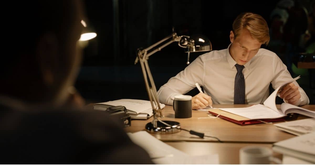 Een privédetective in wit hemd met zwarte das bekijkt een dossier dat op zijn houten bureautafel ligt. Hij heeft een pen in de rechterhand terwijl hij met zijn linkerhand door het dossier bladert. Op tafel: een bureaulamp, zwarte mok, witte mok en andere dossiers. Links vooraan in beeld: het silhouet van de opdrachtgever. 