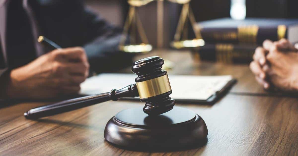 Een gavel of voorzittershamer op een donkere houten tafel. Op de achtergrond: de handen van de rechter (links) en de handen van een schuldeiser (rechts). De rechter heeft een pen in de hand en schrijft op een blad op een klembord.