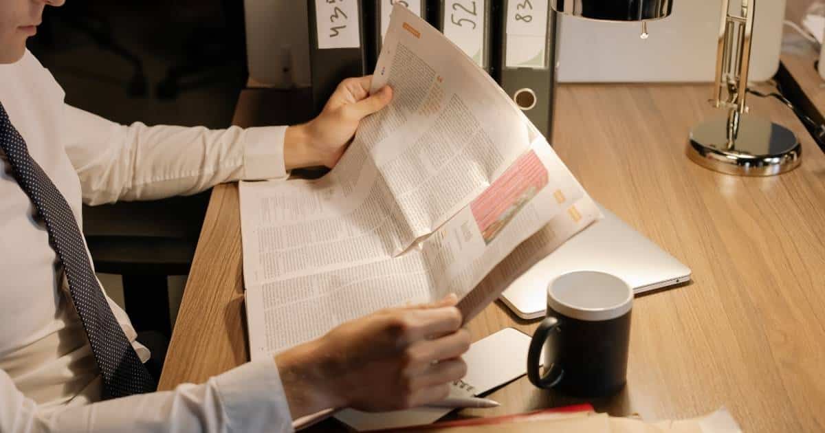 Een privédetective bekijkt een krant. Hij draagt een wit hemd met zwarte das en zit aan een houten bureautafel.