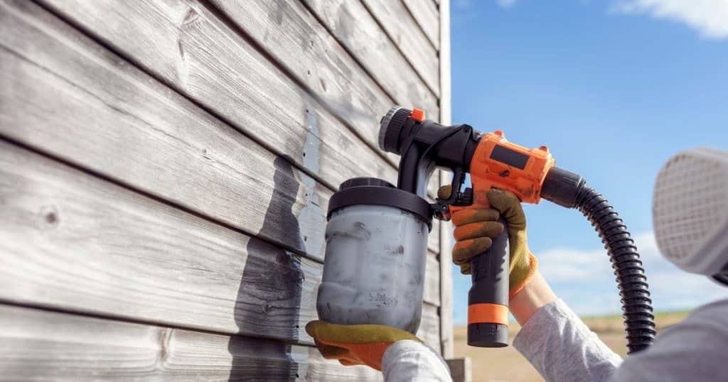 Een schilder spuit grijze verf onder hoge druk op een houten wand. Hij of zij draagt daarbij beschermende handschoenen, mondmasker en kledij.
