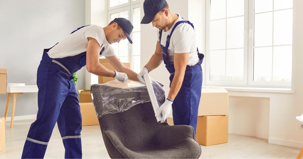 Twee verhuizers in witte t-shirts, blauwe overalls en petjes pakken een designstoel voorzichtig in met folie. Ze staan in een ruimte met witte en grijze wanden, grote ramen en een houten vloer waarop verhuisdozen staan.