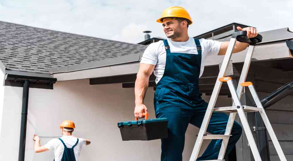 Professionele dakwerker in blauwe overall, en met gele helm en werkkoffer, kruipt op een ladder voor werken aan het dak.