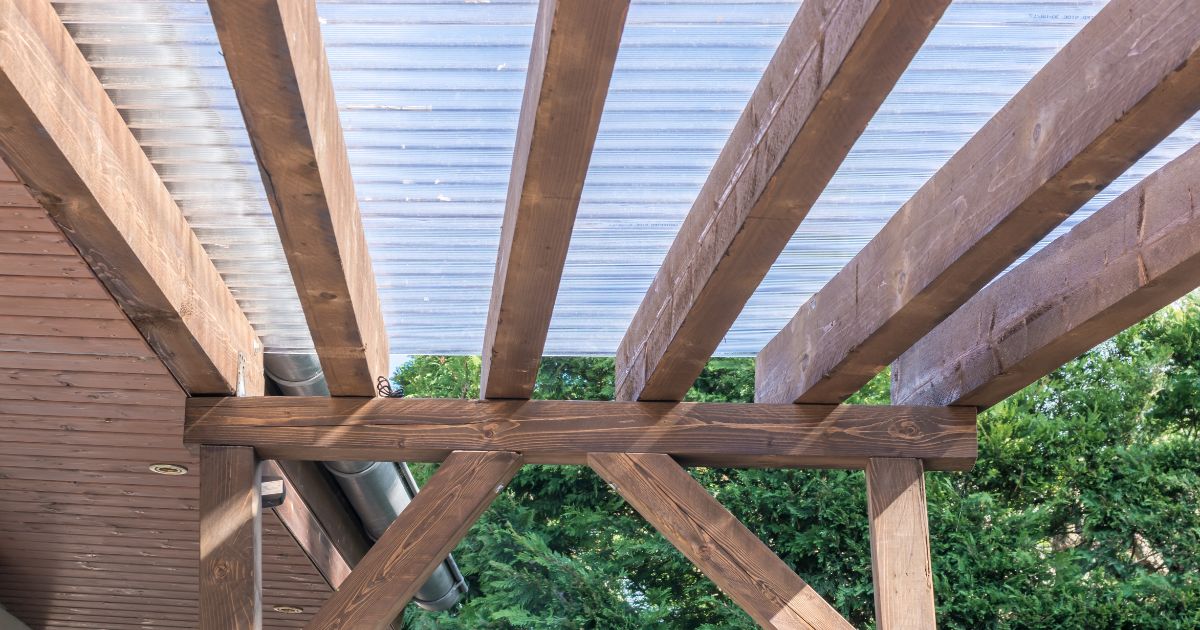 Een veranda met houten balken en een dak uit transparante golfplaten in pvc. Op de achtergrond groen gebladerte.