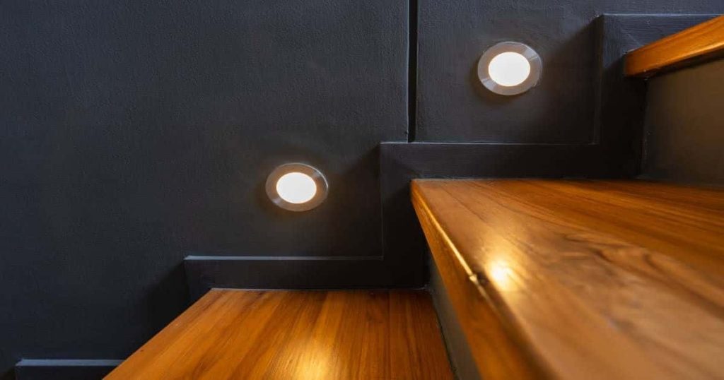 Trap met pvc overzettreden in donkere houtlook en met spots boven elke trede die in de donkergrijze muur zijn geïntegreerd.