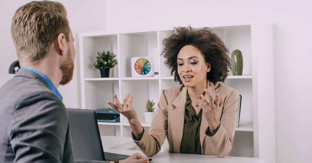 Een dame met krullend haar, een beige blazer en een donkergroen hemd vertelt iets aan de man die voor haar zit. Ze heft haar handen op. De man heeft rossig haar en een baardje, en draagt een donkergrijze blazer boven een blauw hemd. Op de achtergrond een witte muur met daartegen een witte kolomkast met planten, een klok en een typemachine.