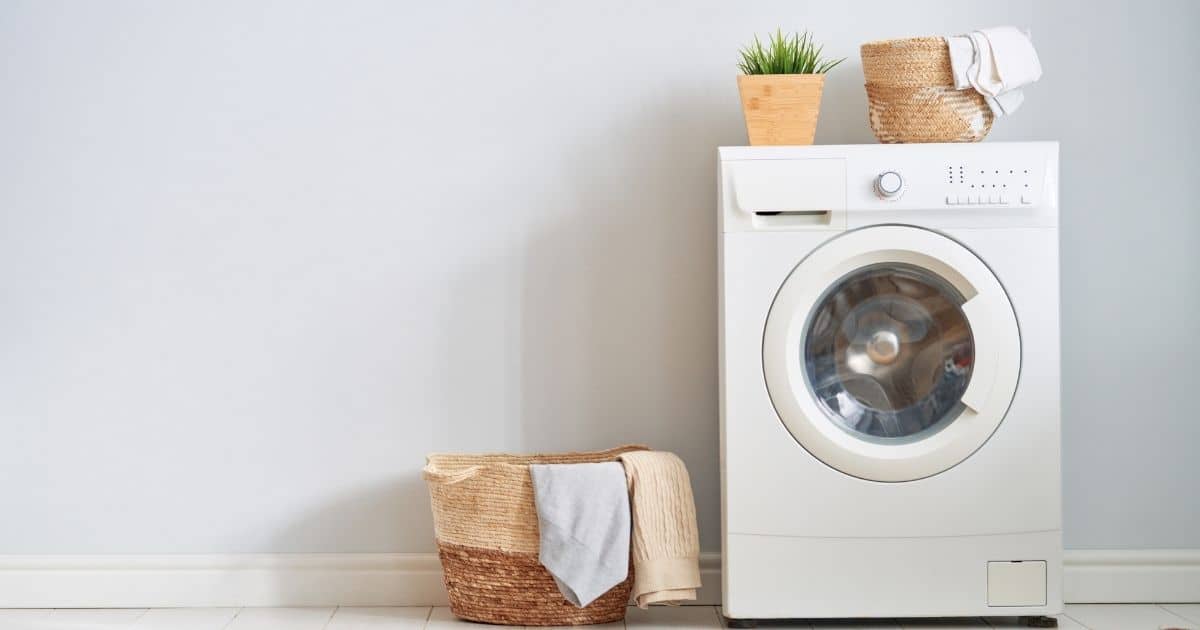 Een witte wasmachine en een rieten wasmand tegen een blauwwitte muur. Op de wasmachine staan een plantje in een houten pot en een rieten mandje met doeken. Over de rand van de rieten wasmand  hangen handdoeken.