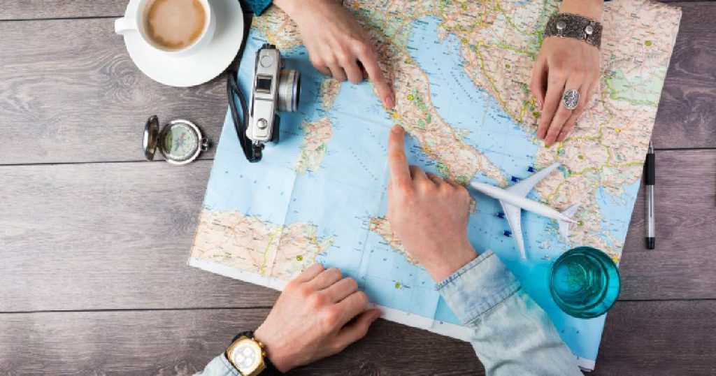 Twee paar handen boven een wereldkaart met een fototoestel, miniatuurvliegtuig, kompas, pen, kopje koffie en glas water.