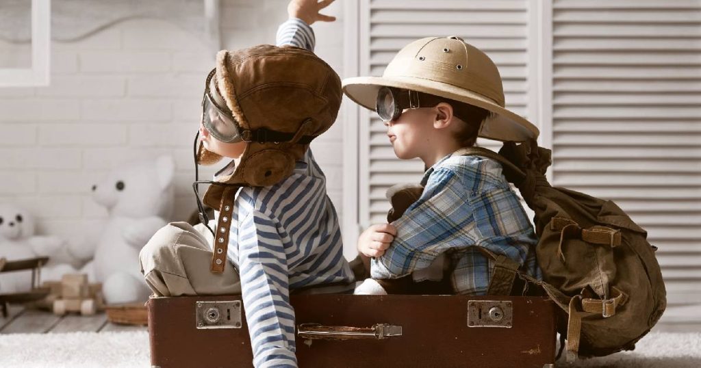 Twee jongetjes in een reiskoffer. Het voorste kind spreidt zijn armen en draagt een vliegeniersbril en -muts. Het achterste jongetje kruist de armen en heeft een rugzak, verkennershoed en vliegeniersbril op.