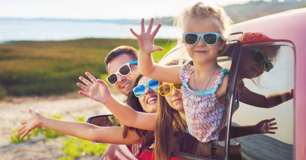 Een moeder, vader en twee dochters met kleurrijke zonnebrillen zwaaien door het raam van een rode auto. 