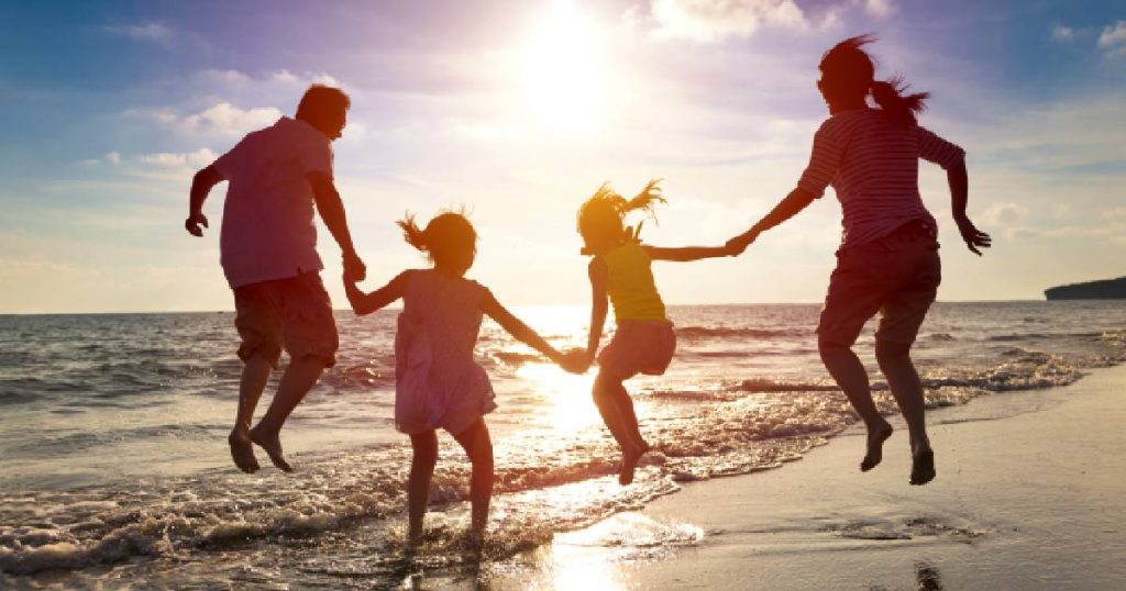 De silhouetten van een moeder, vader en twee dochters op het strand bij zonsondergang - zorgeloos op vakantie dankzij hun reisverzekering.