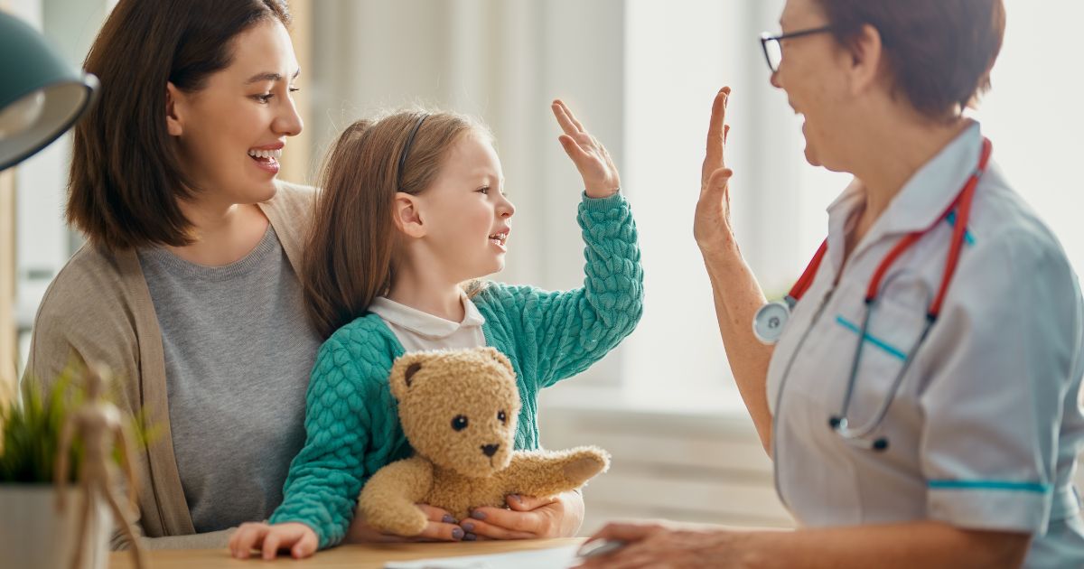 Een moeder en kind op consultatie bij een arts. De arts geeft het kind een high five. Het kind heeft een knuffelbeer bij.