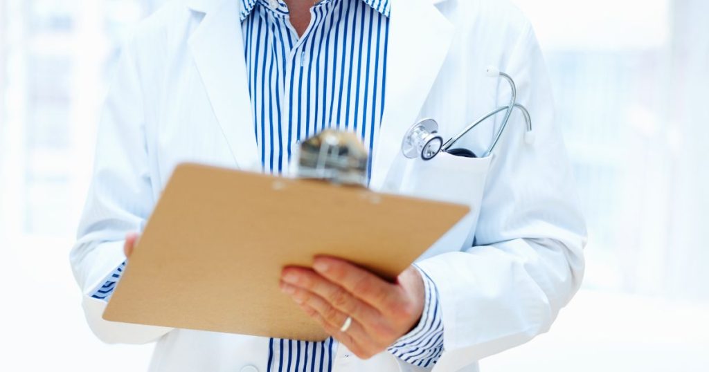 Een dokter met een klembord vult een getuigschrift in. De arts draagt een wit met blauw gestreept hemd, een witte doktersjas en een ring, en heeft een stethoscoop in de borstzak.