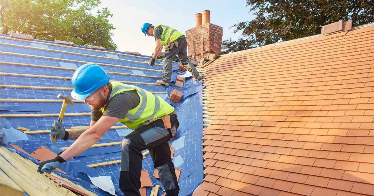 Twee dakwerkers met blauwe veiligheidshelmen en werkkledij renoveren een pannendak. De rechterhelft van het dak verkeert in goede staat. De linkerhelft wordt hersteld: er ligt een nieuwe dakfolie op het dakbeschot, met daarop de tengellatten. 