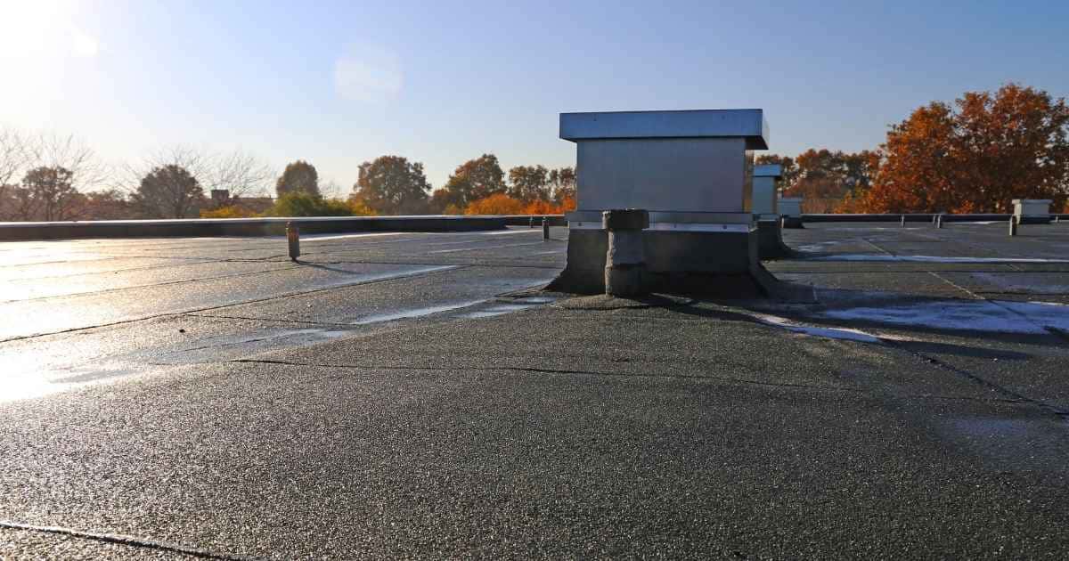 Een nieuw plat dak na renovatie: het dak is bedekt met zwarte dakbedekking met een korrelige structuur. Het materiaal werd in verschillende stroken aangebracht. Op het dak vind je verschillende dakdoorbrekingen voor onder meer ventilatie.