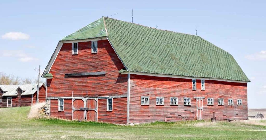 Een rode, houten schuur met een leuk groen schilddak met wolfseind, ook wel een wolfsdak genoemd. De schuur staat op een open grasvlakte.