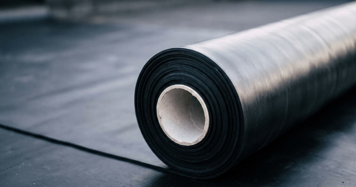 Een opgerolde zwarte EPDM membraan ligt op een donker dakoppervlak met de lichtbruine kartonnen kern zichtbaar in het midden van de rol terwijl het gladde rubber zacht het licht vangt in een onscherpe achtergrond.