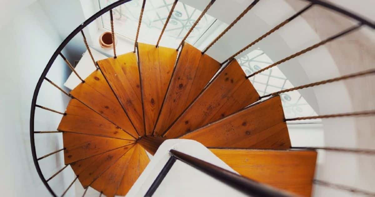 Bovenaanzicht van een spiltrap met een zwarte metalen spil en houten treden, geplaatst in een wit interieur tegen een groot raam.