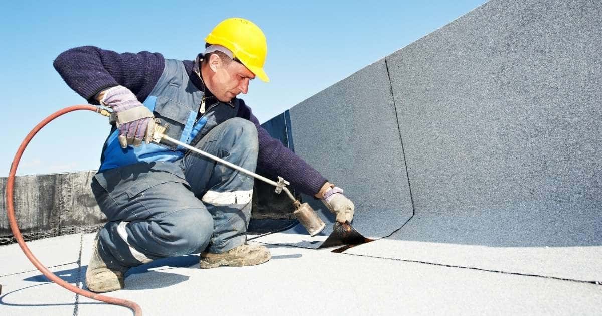 Een dakwerker met gele veiligheidshelm en blauw met grijze werktenue gebruikt een gasbrander om een strook lichtgrijze bitumen op het platte dak te bevestigen.