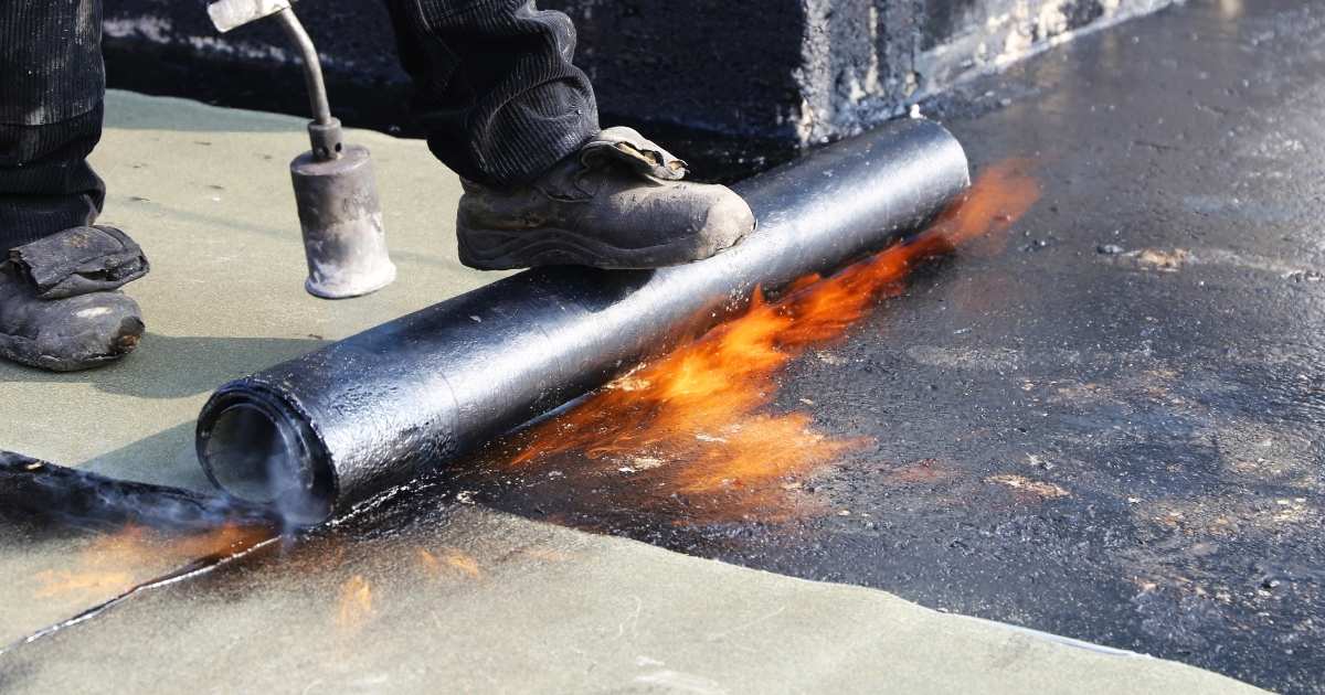 Close-up van een rol roofing of dakleer onder de voet van een dakspecialist. Hij of zij gebruikt een gasbrander om het materiaal op te warmen. De vlammen dansen onder het dakleer.