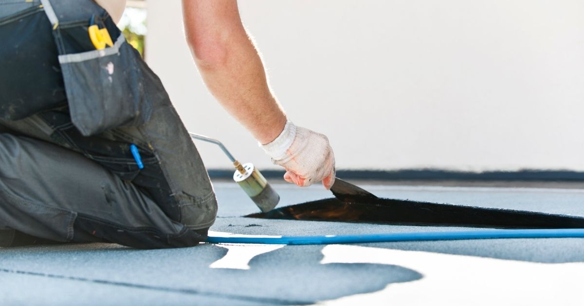 Schuin onderaanzicht van een dakspecialist die een bitumen dak herstelt met een spatel en een gasbrander. 