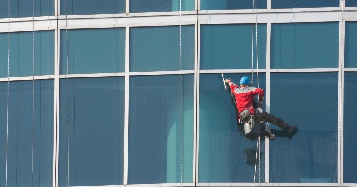 Een ruitenwasser met rode trui, grijze werkbroek en blauwe helm wast de ramen van een appartement. Hij zit op een gondel en gebruikt een trekker om zeep te verwijderen. Aan de gondel hangt en zwarte emmer.
