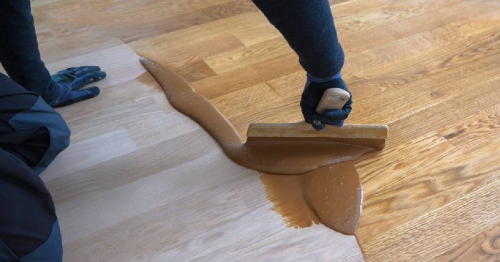 Een persoon in een blauwe overal met blauwe handschoenen werkt een samengesteld parket af met een laag vernis. 