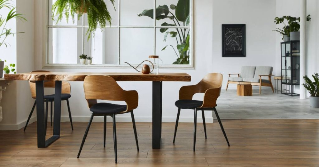 Vooraanzicht van een modern interieur met een samengesteld parket, houten tafel en houten stoelen met ijzeren poten. Op de achtergrond een ruimte met grijze vloer, half afgescheiden van de eerste kamer door een muur met raam. Er staan verschillende kamerplanten in beide ruimtes.