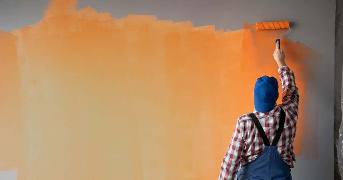 Een man met blauwe pet schildert een grijze muur in het oranje.