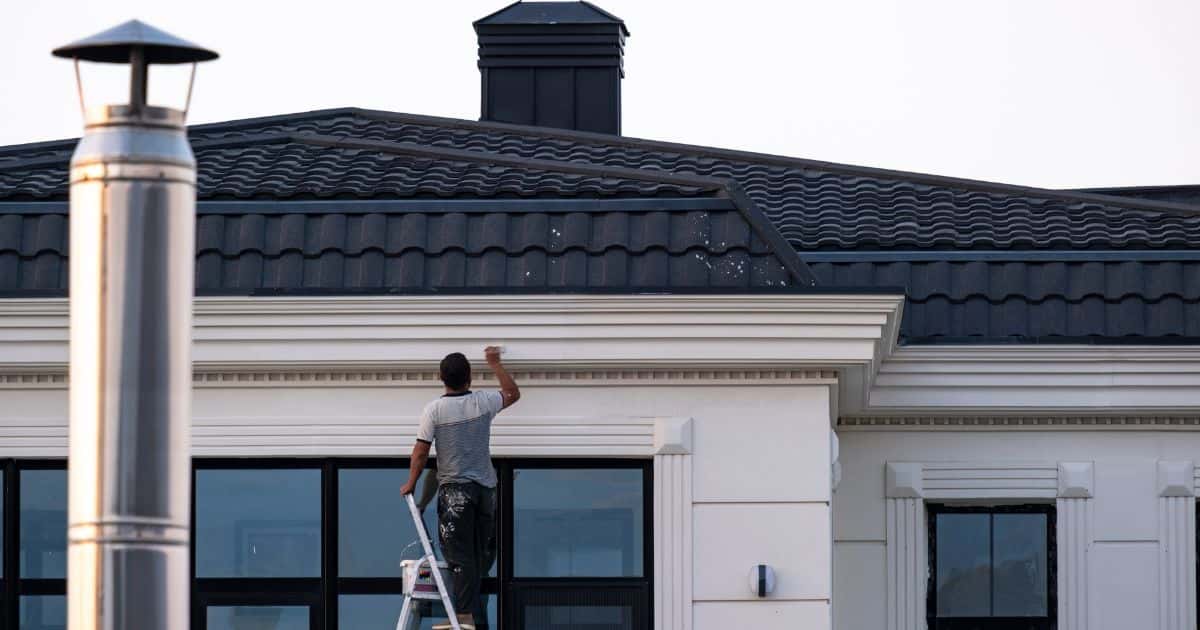een man in halfdonkere kledij schildert de dakgoot van een klassieke woning in het wit. Zo vormt het een mooi geheel met de rest van de woning.