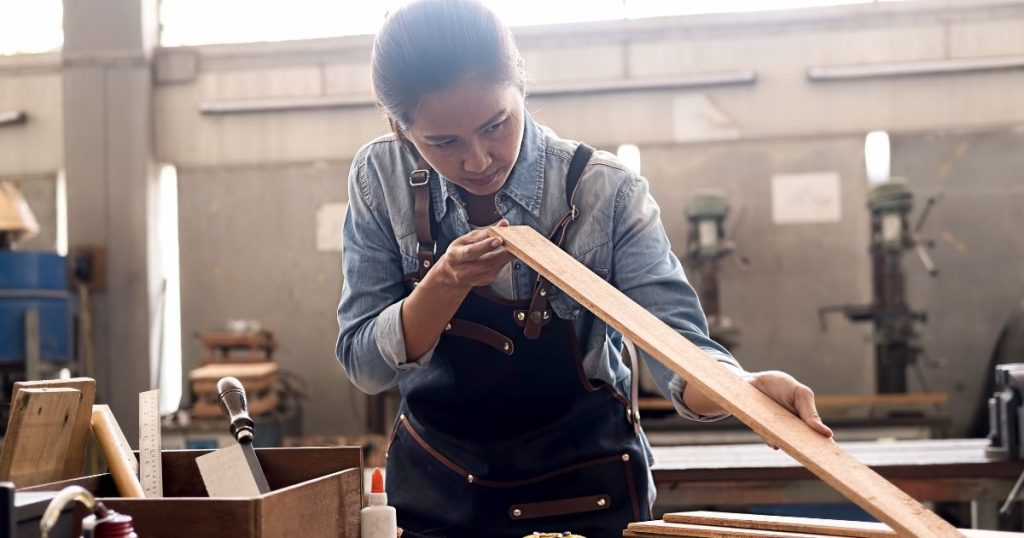 Een vrouw in jeanshemd en grijs-groene overall controleert een houten plank die ze net heeft bewerkt.
