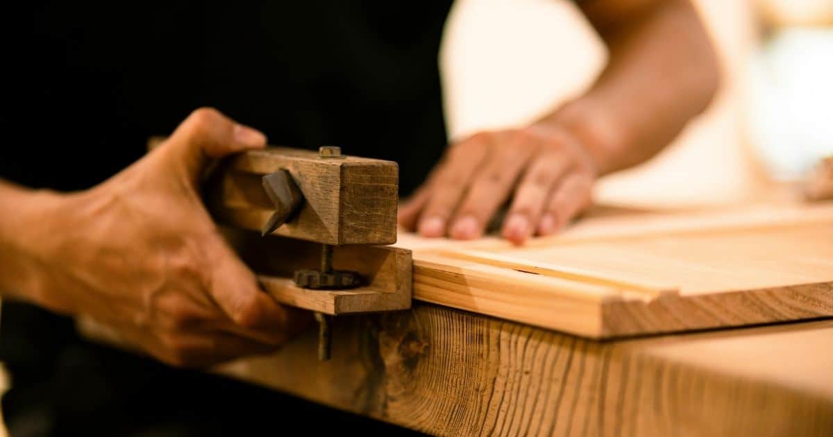 Een schrijnwerker bewerkt massief hout tijdens het bouwen van een kast op maat. Hij houdt met zijn handen houten klem met metalen scharnieren vastL Terwijl de andere hand op een stuk plank rust.