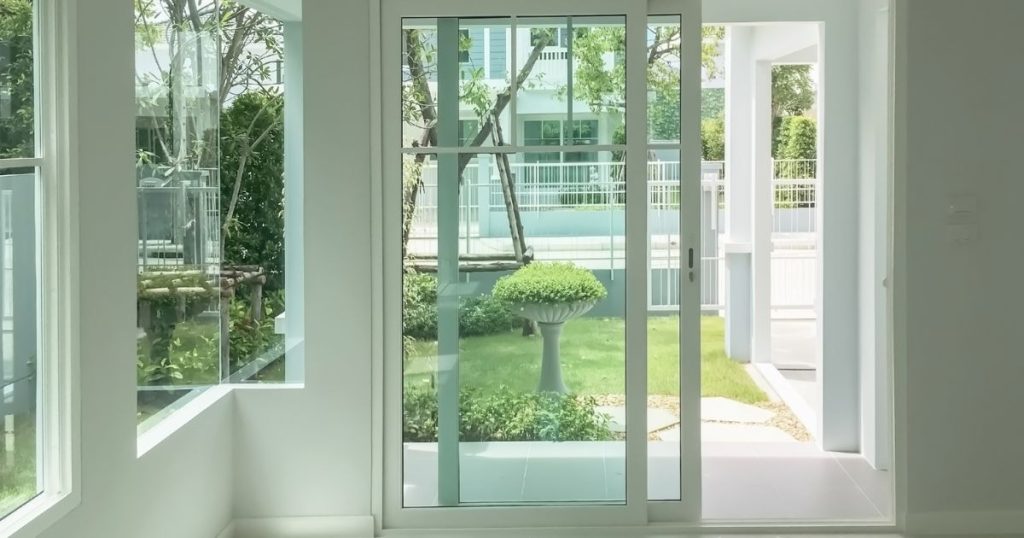 Schuifraam met witte pvc profielen dat vanuit de woonruimte uitgeeft op een overdekt terras en een klein grasperk. De tuin wordt afgeschermd van de straatkant door een wit stalen hek.