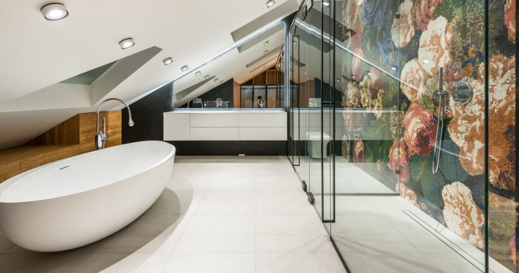 Een design badkamer onder een hellend dak met een ruim bad, een grote wastafel, en een enorme glazen inloopdouche met bloemenwand.