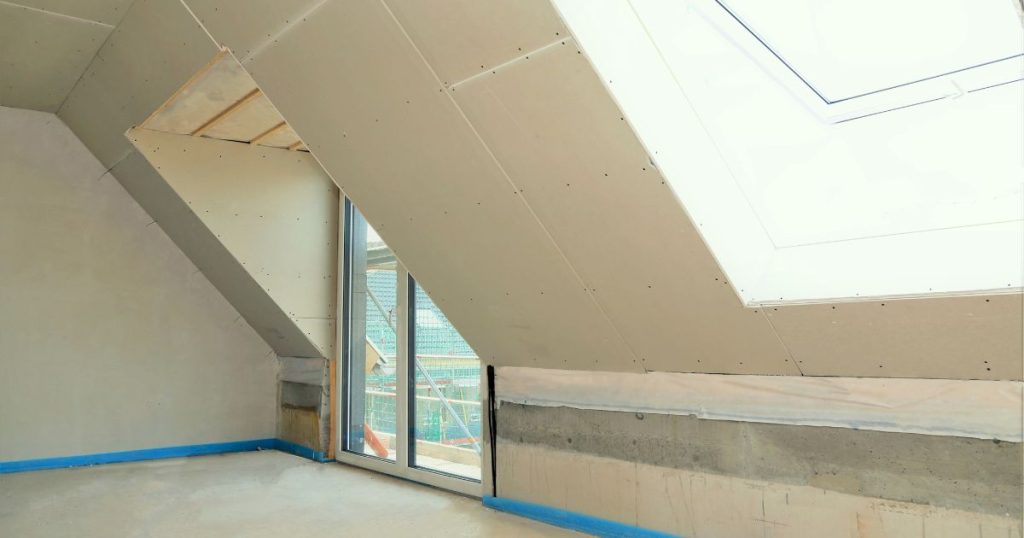 Een recent geïsoleerde zolder waarvan de schuine wanden werden afgewerkt met gipsplaten. Een glazen deur geeft uit op een dakterras. Enkele meters naast de deur zit een dakraam.