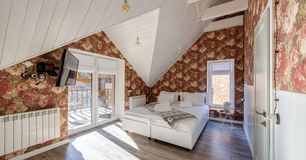 Een afgewerkte zolder die gebruikt wordt als slaapkamer. In de slaapkamer staan een bed en een bijzettafeltje. Een dubbele glazen deur geeft uit op een terras. Naast het bed zit een raam.