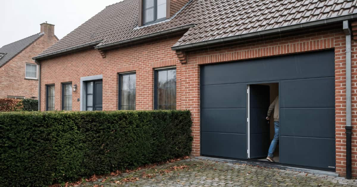 Een bewoner stapt door de geïntegreerde loopdeur in een antracietgrijze sectionaalpoort aan een Vlaamse bakstenen woning met kasseioprit en groene haag op een bewolkte dag.