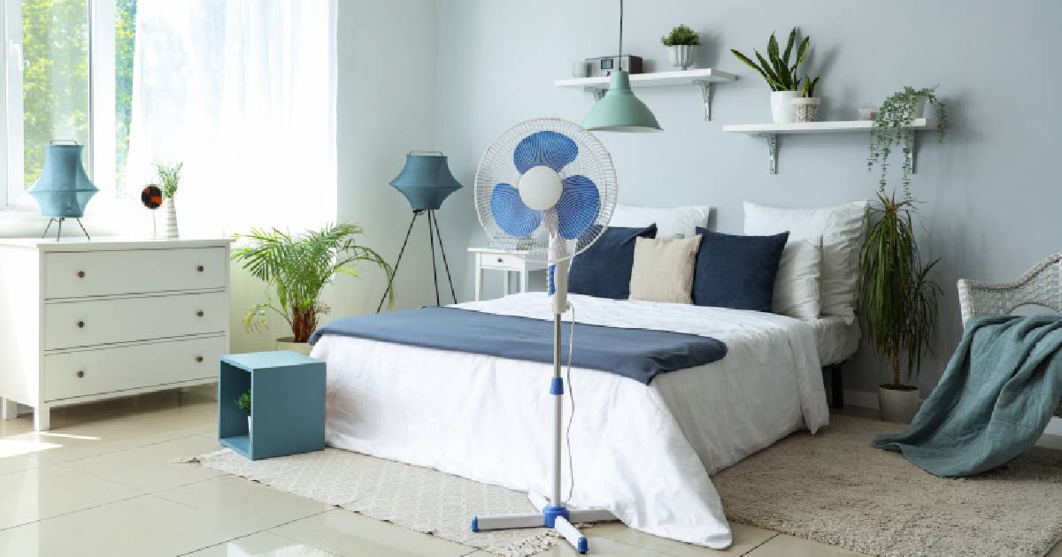 Een slaapkamer zonder airco die men wilt koelen met een ventilator. In de slaapkamer staat een bed en een kast, op de tegelvloer liggen twee tapijten en aan de licht blauwgrijze wand hangen witte wandplanken. In de kamer staan verschillende kamerplanten die ook bijdragen tot een aangename slaaptemperatuur.