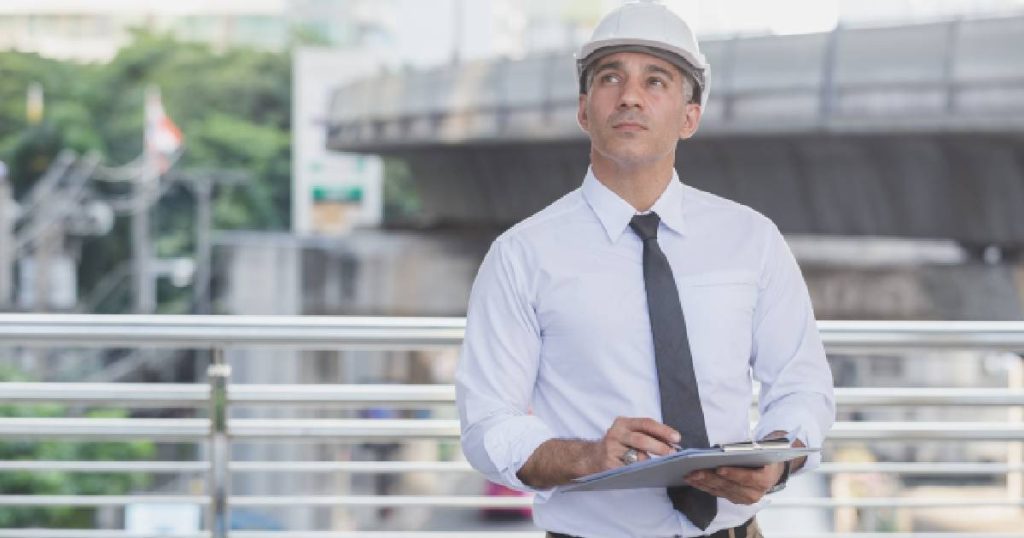 Een sloopdeskundige van Tracimat in een wit hemd met grijze das en witte helm houdt een klembord met conform verklaard sloopopvolgingsplan vast.