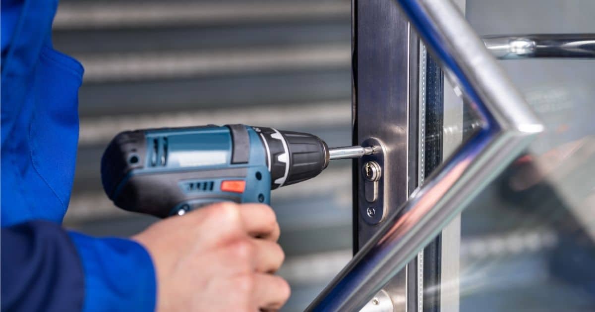 Een slotenmaker plaatst een nieuw slot in een ijzeren deur. Het gaat om een insteekslot. Enkel zijn hand en arm zijn in beeld, alsook de boor waarmee hij het slot vastmaakt in de deur.