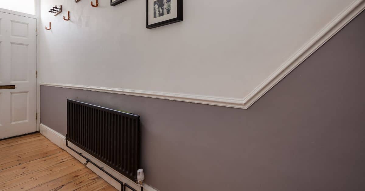 Een muur in een hal in wit en grijs, waarbij een detail van de (oude) lambrisering de overgang vormt tussen beide delen. De zwarte chauffage en andere decoratie geven het geheel een tijdloze uitstraling.