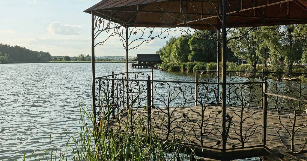 Een open, vrijstaande smeedijzeren veranda aan de oever van een meer. Het smeedijzer is verwerkt tot natuurlijke, krullende vormen.