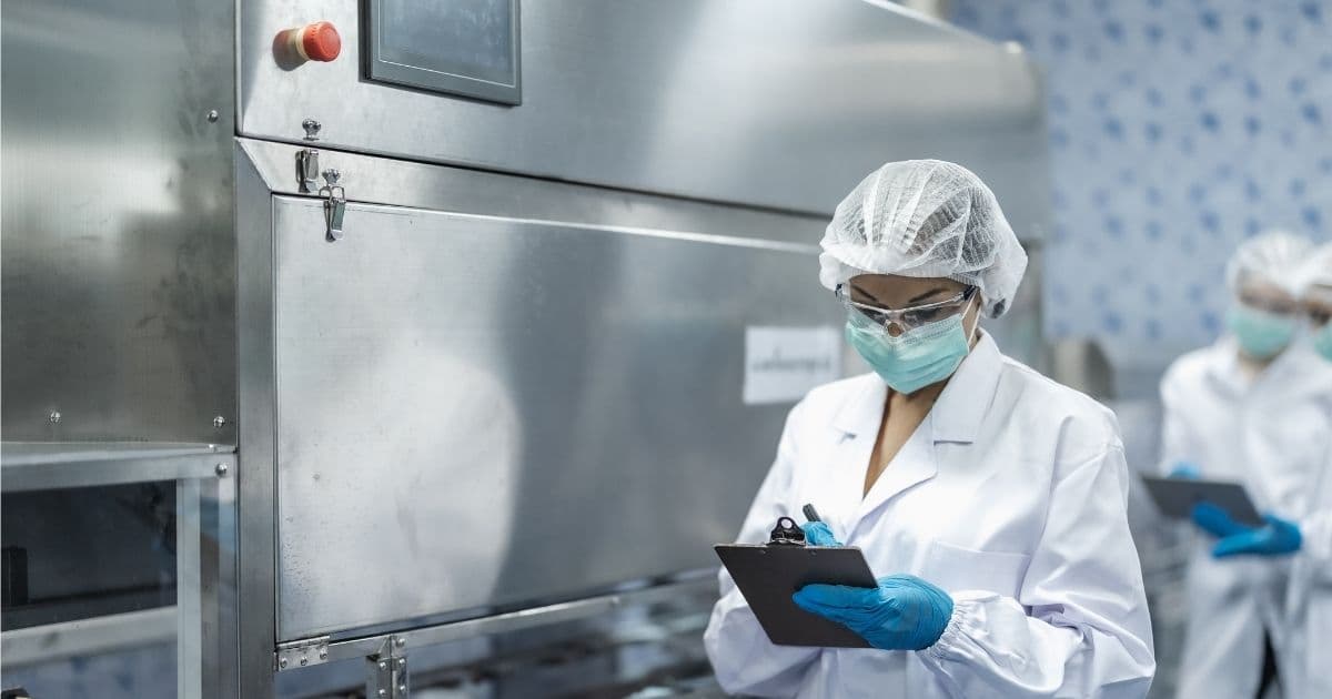Een lid van een dienst voor bescherming en preventie op het werk controleert een metalen installatie in een bedrijf. Ze draagt een lichtgroen mondmasker, semi-transparant haarnet, een witte jas en blauwe hygiënehandschoenen. Ze heeft een klembord vast waarop ze iets noteert.