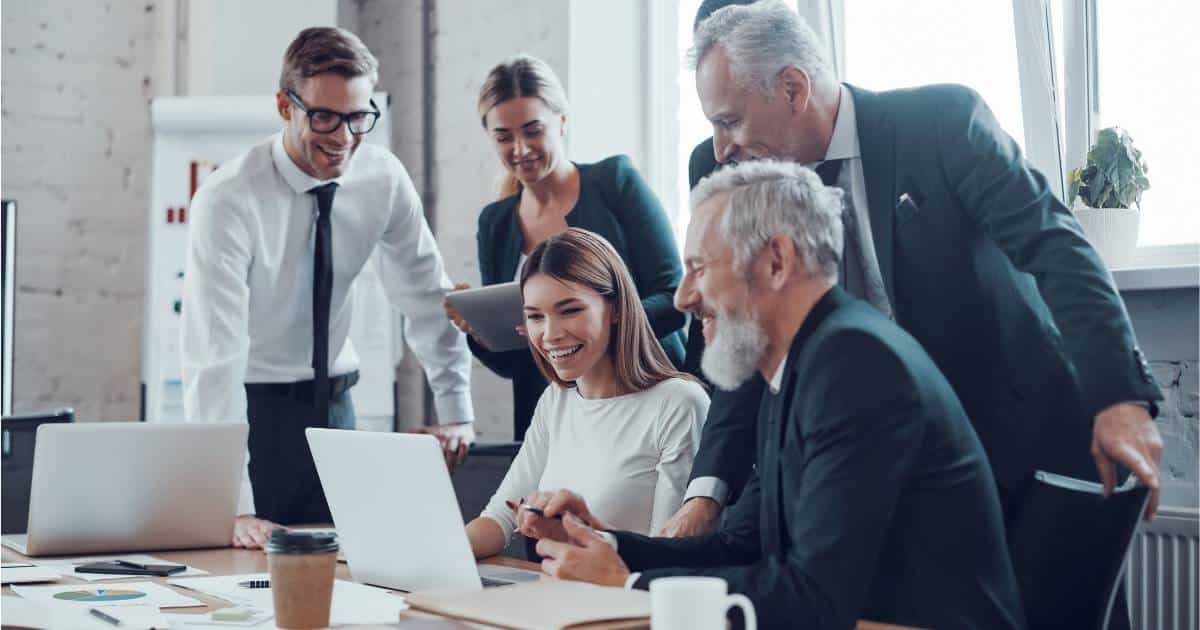 Een groep van 6 collega's verzamelt zich rond een witte laptop. Ze dragen werkkledij. De voorste twee zijn mannen met wit haar. Het dichtst bij de laptop zit een jonge vrouw met lang bruin haar en een wit t-shirt. Rechts achter haar staat een vrouw met lang lichtbruin haar en een donkerblauwe blazer: ze heeft een laptop of tablet vast. Naast de dame staat een man met bril, wit hemd, zwarte das en zwarte broek. De zesde persoon is niet duidelijk in beeld: hij staat achter één van de witharige mannen.