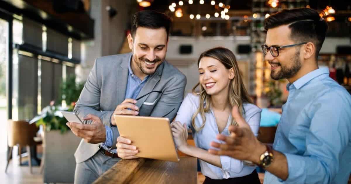 Drie personen staan in een open ruimte aan een hoge tafel. Ze bekijken iets op een goudkleurige tablet. De man in grijze kostuum houdt ook een bril en smartphone vast.