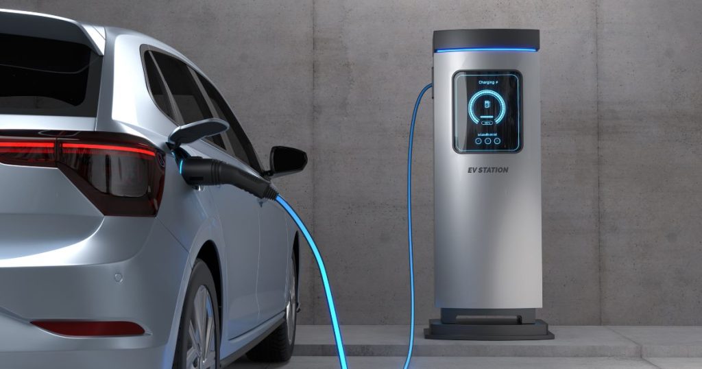 Een lichtgrijsblauwe elektrische auto laadt op via een laadpaal in een grijze betonnen garage.