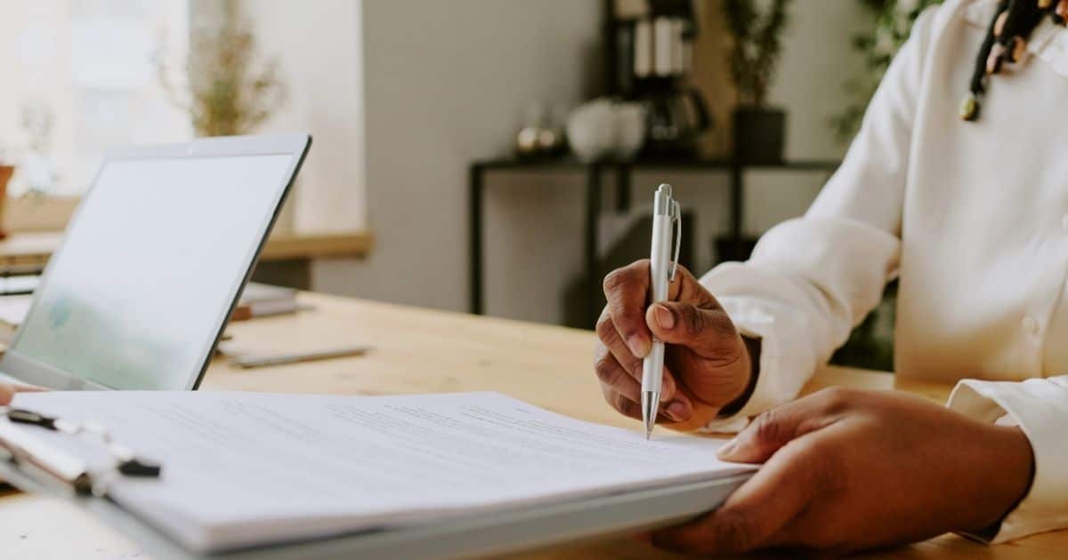 Een dame in wit hemd houdt een pen boven een contract in een wit klembord. Ze zit aan een lichte houten tafel, waarop ook een laptop staat.